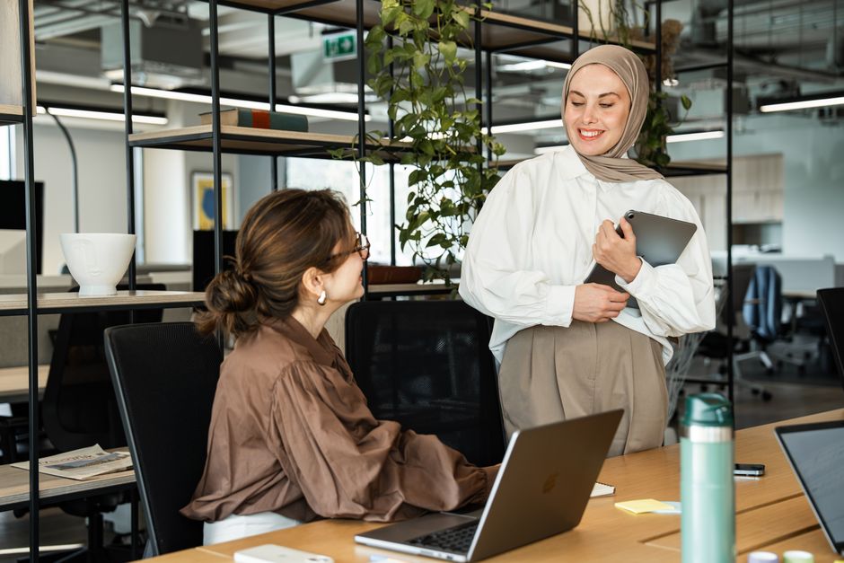 Two professional women in a modern office setting engaging in a positive interaction using laptops.